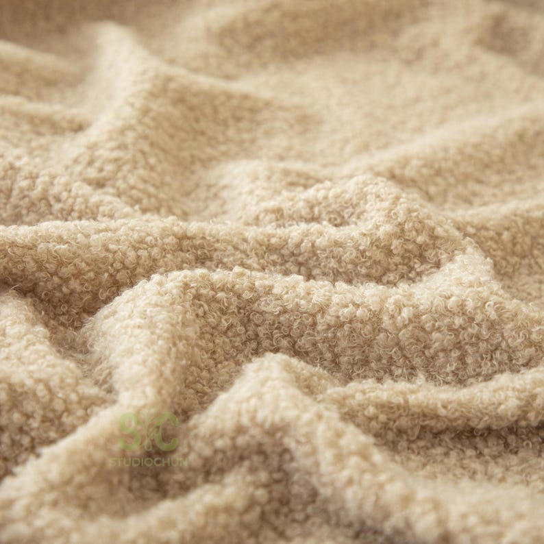 Op de afbeelding: Een beige stof met een dik, krullend en getextureerd oppervlak. Het materiaal vertoont diepe plooien en rimpelingen, wat zorgt voor een zachte en dichte uitstraling. Een klein lichtgroen logo met de tekst STUDIOCHUN is linksonder zichtbaar.