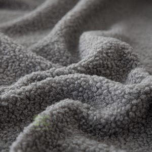Op de afbeelding: Een grijze stof met een dicht, krullend en getextureerd oppervlak. Het materiaal vertoont diepe plooien en rimpelingen, wat zorgt voor een zacht en onregelmatig uiterlijk. Een klein, vaag logo met de tekst STUDIOCHUN is linksonder zichtbaar.