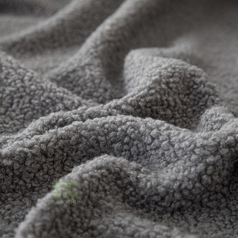 Op de afbeelding: Een grijze stof met een dicht, krullend en getextureerd oppervlak. Het materiaal vertoont diepe plooien en rimpelingen, wat zorgt voor een zacht en onregelmatig uiterlijk. Een klein, vaag logo met de tekst STUDIOCHUN is linksonder zichtbaar.