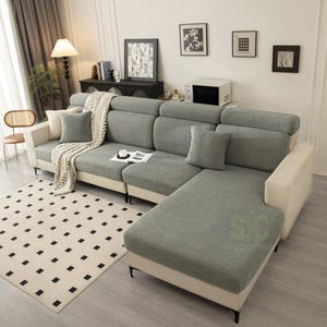 May include: A sectional sofa featuring a textured, sage green slipcover over the seat and back cushions, contrasted by a solid cream-colored frame. The sofa includes matching sage green throw pillows and a cream-colored throw blanket draped over one arm.