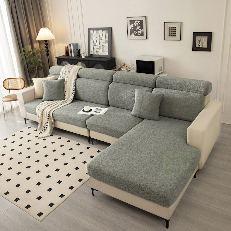 May include: A sectional sofa featuring a textured, sage green slipcover over the seat and back cushions, contrasted by a solid cream-colored frame. The sofa includes matching sage green throw pillows and a cream-colored throw blanket draped over one arm.