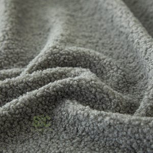 Op de afbeelding: Een grijze stof met een dicht, krullend en getextureerd oppervlak. Het materiaal vertoont diepe plooien en rimpelingen die een spel van licht en schaduw op het oppervlak cre&euml;ren. Een klein, groen, gestileerd logo met de letters SC is zichtbaar.