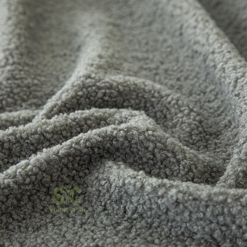 Op de afbeelding: Een grijze stof met een dicht, krullend en getextureerd oppervlak. Het materiaal vertoont diepe plooien en rimpelingen die een spel van licht en schaduw op het oppervlak cre&euml;ren. Een klein, groen, gestileerd logo met de letters SC is zichtbaar.