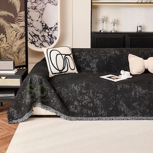 Puede incluir: Una funda de sofá negra texturizada con un borde con flecos está colocada sobre un sofá blanco. Cojines decorativos en crema y negro con diseños abstractos y forma de lazo están colocados en el sofá. Se ve una pequeña mesa con un altavoz.
