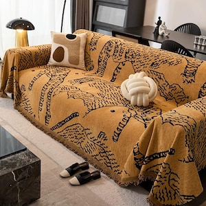 May include: A mustard-colored sofa throw with a black animal print design. The throw is draped over a sofa with a matching pillow and a white knot-shaped decorative object. Black and white shoes are placed on the floor.