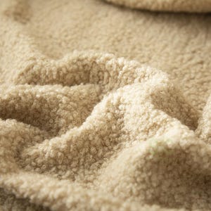 May include: A beige fabric with a dense, curly, sherpa-like texture. The material features a thick, looped pile surface that creates deep folds and shadows across the frame, highlighting the soft, fuzzy, and voluminous nature of the textile.