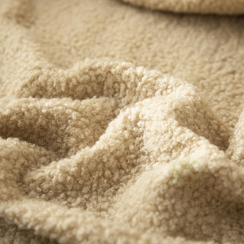 May include: A beige fabric with a dense, curly, sherpa-like texture. The material features a thick, looped pile surface that creates deep folds and shadows across the frame, highlighting the soft, fuzzy, and voluminous nature of the textile.