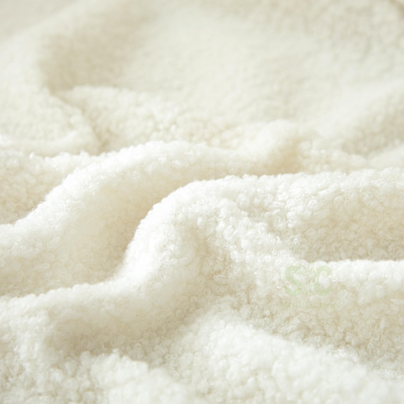 May include: A close-up view of off-white, textured sherpa fleece fabric with a soft, curly pile. The material features deep folds and ripples, creating a sense of depth and volume. A small, faint logo is visible in the lower right corner.