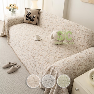 May include: Beige floral patterned sofa cover with fringe trim. A decorative pillow with a floral design and a white teacup on a saucer are on the sofa. A pair of beige slippers are on the floor.