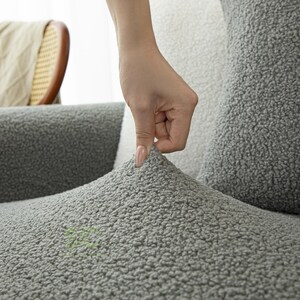 Op de afbeelding: Een grijze, gestructureerde bankhoes gemaakt van een zacht, fleece-achtig materiaal met een dikke, noppige pool. De stof wordt omhoog getrokken, wat de flexibele en rekbare aard aantoont terwijl het het oppervlak van het meubelstuk bedekt.