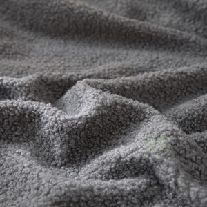 May include: A close-up view of a grey fabric with a thick, curly, and textured pile. The material features a dense, looped surface that creates deep folds and shadows across the frame, highlighting the soft and fuzzy nature of the textile.