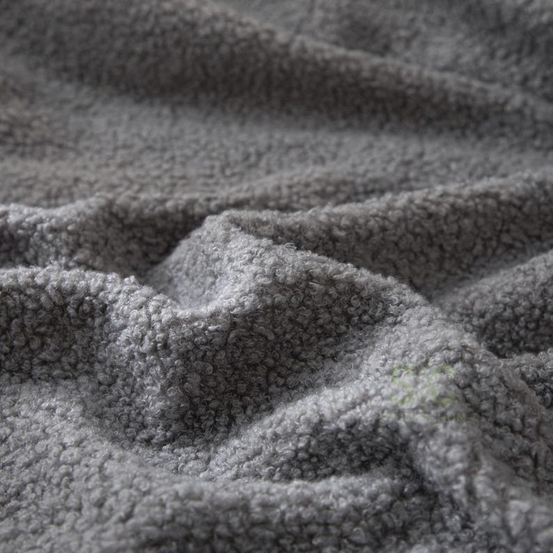 May include: A close-up view of a grey fabric with a thick, curly, and textured pile. The material features a dense, looped surface that creates deep folds and shadows across the frame, highlighting the soft and fuzzy nature of the textile.