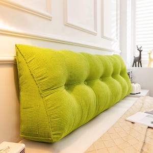 Puede incluir: Una almohada triangular de respaldo de pana verde lima. La almohada tiene una serie de secciones redondeadas grandes y abotonadas. La almohada se coloca contra una pared blanca y una cama con una manta de color crema.