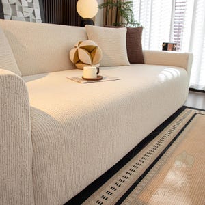 Peut inclure: Un canapé crème avec un tissu texturé, orné de coussins décoratifs beiges et bruns. Un tapis à motifs avec des bordures noires et une tasse à café sur une petite table complètent le cadre du salon.