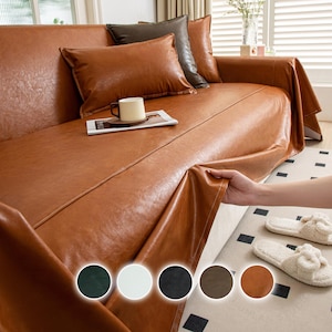 May include: A brown leather sofa cover draped over a sofa, with matching cushions. The cover is being held up by a hand, revealing the sofa underneath. A cup and magazine rest on the sofa. Colour options are shown at the bottom.