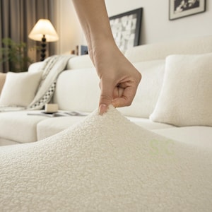 May include: A cream-colored, textured sherpa fabric cover draped over a sofa. The material features a dense, curly, fleece-like pile with a soft, plush appearance. The fabric is pulled upward to demonstrate its stretch and thick, fuzzy surface texture.