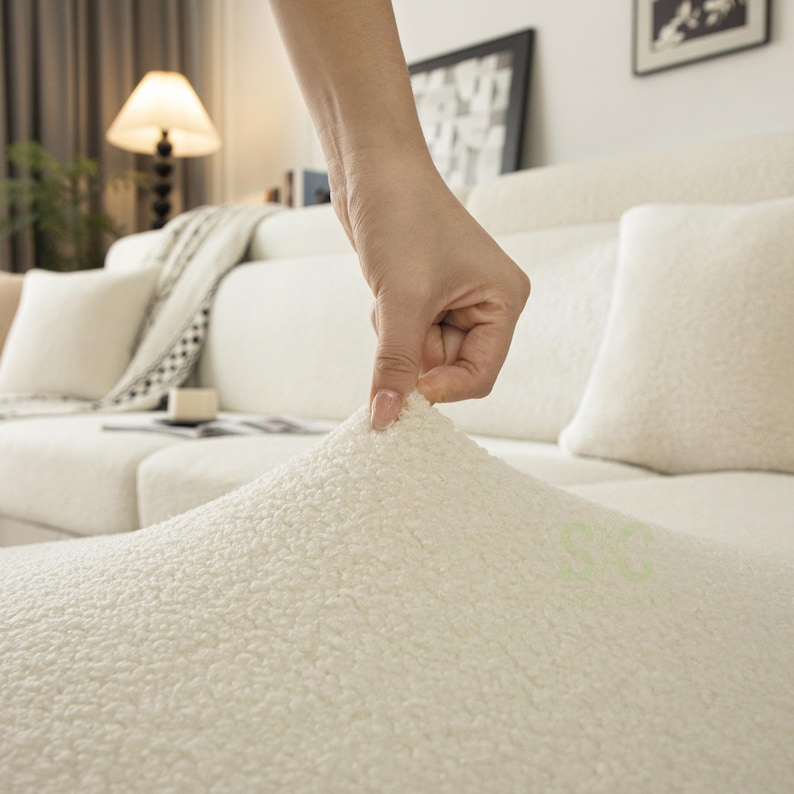 May include: A cream-colored, textured sherpa fabric cover draped over a sofa. The material features a dense, curly, fleece-like pile with a soft, plush appearance. The fabric is pulled upward to demonstrate its stretch and thick, fuzzy surface texture.