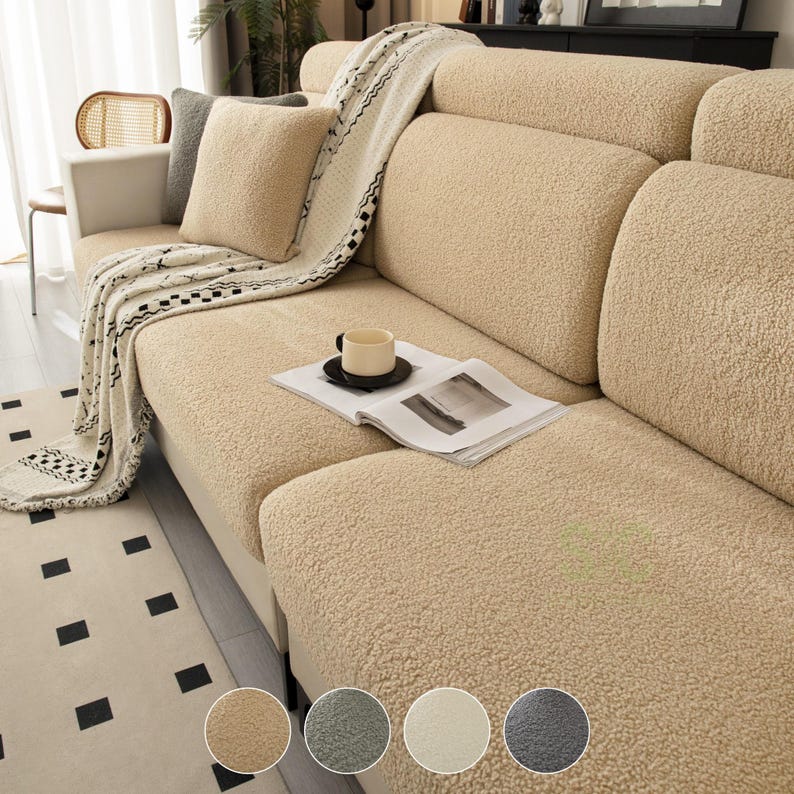 May include: A beige sherpa-style sofa cover with a textured, fleece-like surface. The cover is draped over a sofa, accompanied by a matching beige throw blanket, a beige throw pillow, an open book, and a small black mug on the seat cushion.