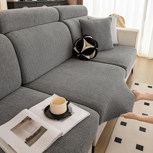 Puede incluir: Un sofá seccional gris con una tela texturizada. Una bola decorativa en blanco y negro y un cojín gris descansan sobre el sofá. Una taza de café está sobre una revista. El sofá se está ajustando.