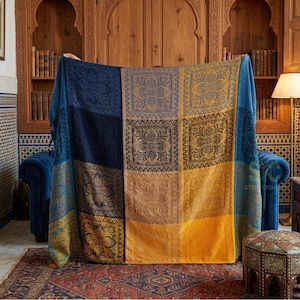 Könnte beinhalten: Eine große, farbenfrohe Decke mit einem geometrischen Muster. Die Decke zeigt Quadrate in Marineblau, Gold und Beige mit aufwendigen floralen Designs. Sie ist über einem blauen Sofa in einem Raum mit einem Holzschrank und einem gemusterten Teppich drapiert.