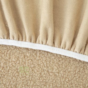 Op de afbeelding: Een beige stoffen hoes met een geplooide, gerimpelde textuur langs de bovenrand, vastgezet met een witte elastische band. Het onderste gedeelte van het item bestaat uit een dicht, gestructureerd, krullend beige materiaal met een zachte uitstraling.