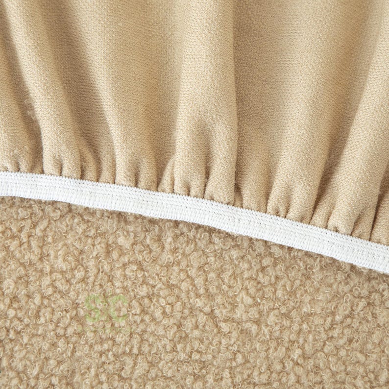 Op de afbeelding: Een beige stoffen hoes met een geplooide, gerimpelde textuur langs de bovenrand, vastgezet met een witte elastische band. Het onderste gedeelte van het item bestaat uit een dicht, gestructureerd, krullend beige materiaal met een zachte uitstraling.