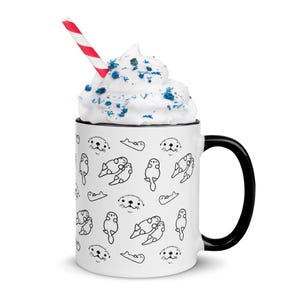 May include: White ceramic mug with a black handle and rim, featuring a repeating pattern of black line-drawn otters. The mug is filled with whipped cream and blue sprinkles, and a red and white striped straw.