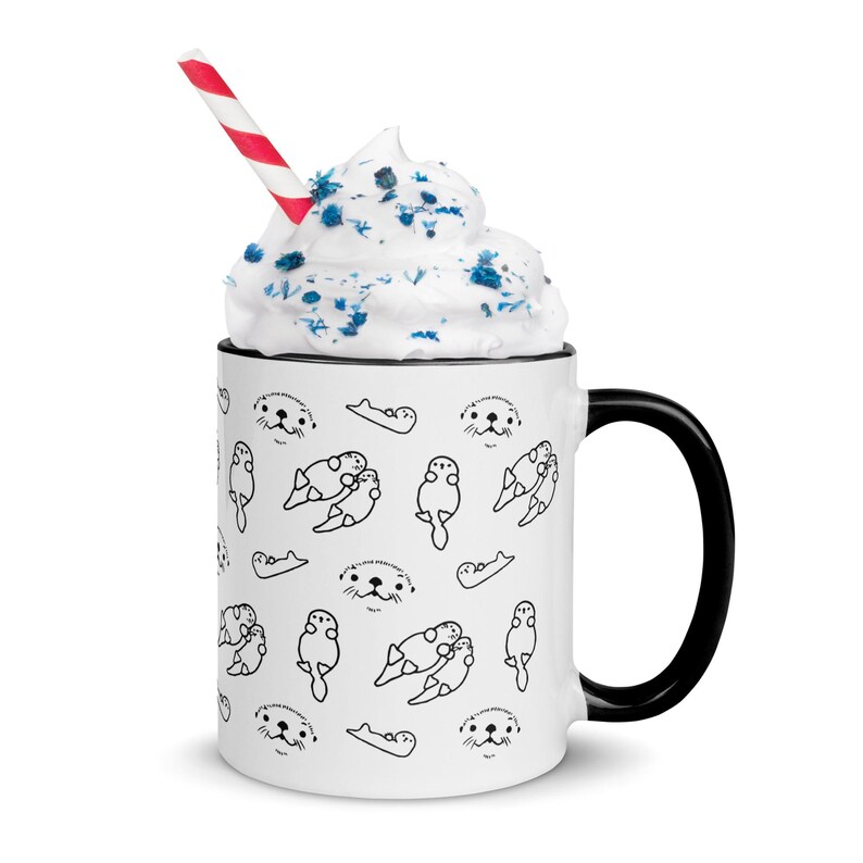 May include: White ceramic mug with a black handle and rim, featuring a repeating pattern of black line-drawn otters. The mug is filled with whipped cream and blue sprinkles, and a red and white striped straw.