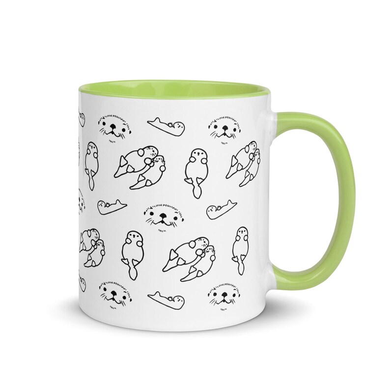 May include: White ceramic mug with a light green interior and handle. The mug features a repeating pattern of black line drawings of otters in various poses. The otters are depicted swimming, cuddling, and with their faces visible, creating a playful design.