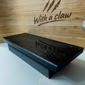 Puede incluir: Una tabla de cortar rectangular negra con una superficie texturizada se asienta sobre una base gris oscuro. El fondo presenta una superficie de madera con el texto "With a claw" y garras.