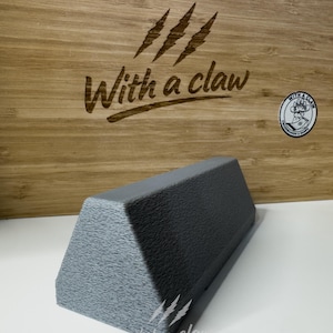 Puede incluir: Un objeto triangular gris texturizado con una base plana. El objeto está frente a una superficie de madera con el texto "With a claw" y marcas de garras grabadas. Una pegatina con el mismo texto es visible.