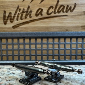 May include: Two silver skateboard trucks are displayed on a speckled surface. The trucks are in the foreground, with a wooden background featuring claw marks and the text "With a claw". The trucks have a metallic finish.
