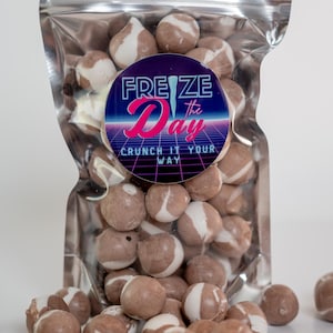 May include: A clear, metallic bag filled with round, brown candies swirled with white. A circular sticker on the bag reads "FREZE the Day" in blue and pink, with the tagline "CRUNCH IT YOUR WAY". Some candies are scattered outside the bag.
