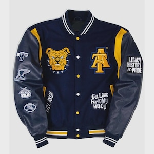 Könnte beinhalten: Marineblaue und goldfarbene Collegejacke mit Lederärmeln. Mit einem Bulldoggen-Patch, den Buchstaben "A" und "T" sowie dem Text "Legacy History Pride" und "Got Love For My HBCU". Die Jacke hat weiße Streifen an Kragen, Bündchen und Saum.