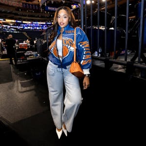 May include: A blue and orange New York Knicks bomber jacket with a basketball graphic, worn over a white crop top with light-wash wide-leg jeans, white pointed-toe heels, and a tan shoulder bag with a long strap, captured in a stadium setting.