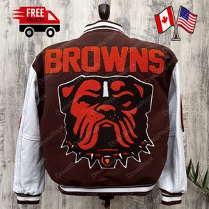 May include: Brown varsity jacket with white sleeves. The back features the word "BROWNS" in orange lettering above a bulldog logo. The jacket has a white collar and cuffs with a striped design. A classic athletic style.