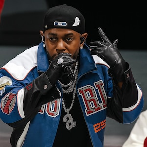 Puede incluir: Una persona con una gorra de béisbol negra, una chaqueta azul y blanca y guantes negros. La chaqueta tiene letras y parches rojos y blancos. La persona lleva joyas de diamantes y sostiene un micrófono.