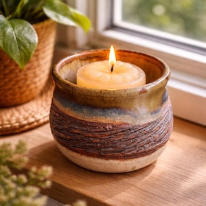 May include: A lit candle in a ceramic holder sits on a wooden surface. The candle is a light yellow color and the holder has a textured, earthy design with brown, blue, and tan tones. A potted plant is in the background.