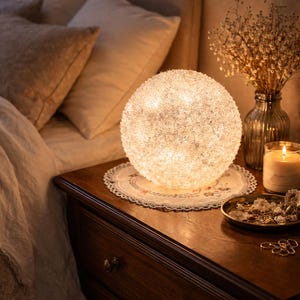 May include: A glowing, spherical decorative lamp with a textured surface, resting on a lace doily atop a dark wooden nightstand. A lit candle, a small dish of jewelry, and a vase of dried flowers complete the cozy bedroom scene.
