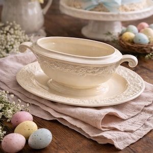 May include: Cream-colored gravy boat and saucer set with embossed floral detailing. The set sits on a pink linen napkin, with pastel Easter eggs nearby. A cake with blue ribbon and a basket of eggs are in the background.