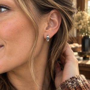 May include: A small silver hoop earring featuring a single round, dark blue gemstone set in the centre. The earring has a polished metallic finish and a hinged closure, worn on the earlobe to provide a subtle and reflective appearance.