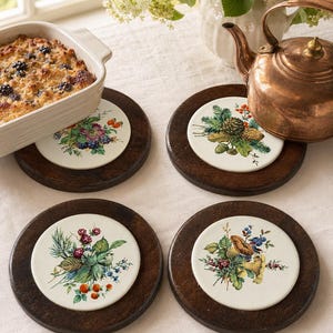 May include: A set of four circular trivets featuring dark brown wooden frames and white ceramic centers. Each ceramic insert displays a unique botanical illustration of berries, pinecones, mushrooms, or forest foliage in vibrant colors.