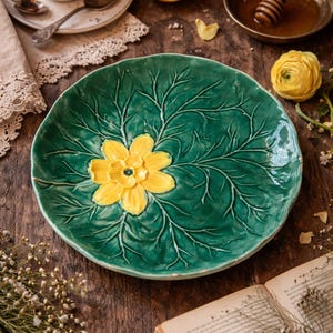 May include: A green ceramic plate with a leaf design and a yellow flower in the center. The plate is on a wooden surface, with a cup of tea, lemon, honey, and a book in the background.