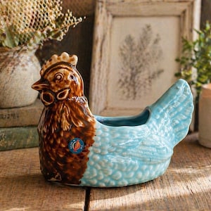 May include: A ceramic chicken-shaped planter with a brown head and body, and a light blue tail. The planter has a small opening for plants and a decorative design. A small sticker is on the side.