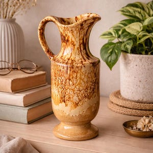 May include: A ceramic pitcher featuring a glossy finish with a mottled brown and cream glaze. The vessel has a curved handle, a wide pouring spout, and a pedestal base, showcasing a distinct drip pattern that transitions from dark brown to light tan.