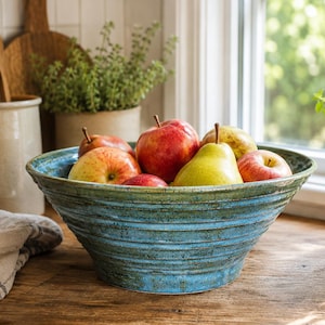 May include: A blue ceramic bowl filled with fresh apples and a pear. The bowl has a textured, ridged design and sits on a wooden surface. Kitchen utensils and plants are visible in the background, near a window.