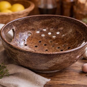 May include: A brown ceramic colander with drainage holes and two handles. The colander sits on a wooden surface with garlic, thyme, and a linen cloth. A basket of lemons is in the background.