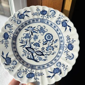 May include: A white ceramic plate with a blue floral design. The plate has a scalloped edge and features a central floral motif surrounded by smaller floral and fruit patterns. The plate is approximately 8 inches in diameter.