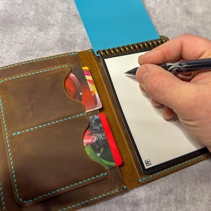 May include: Brown leather notebook and wallet combination with turquoise stitching. The wallet section has card slots with colorful cards visible. A person is writing on the notepad with a black pen.