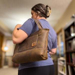 May include: A brown leather tote bag with a front pocket and a handle. The bag has a rectangular shape with visible stitching and a decorative emblem. The bag is being carried over the shoulder.
